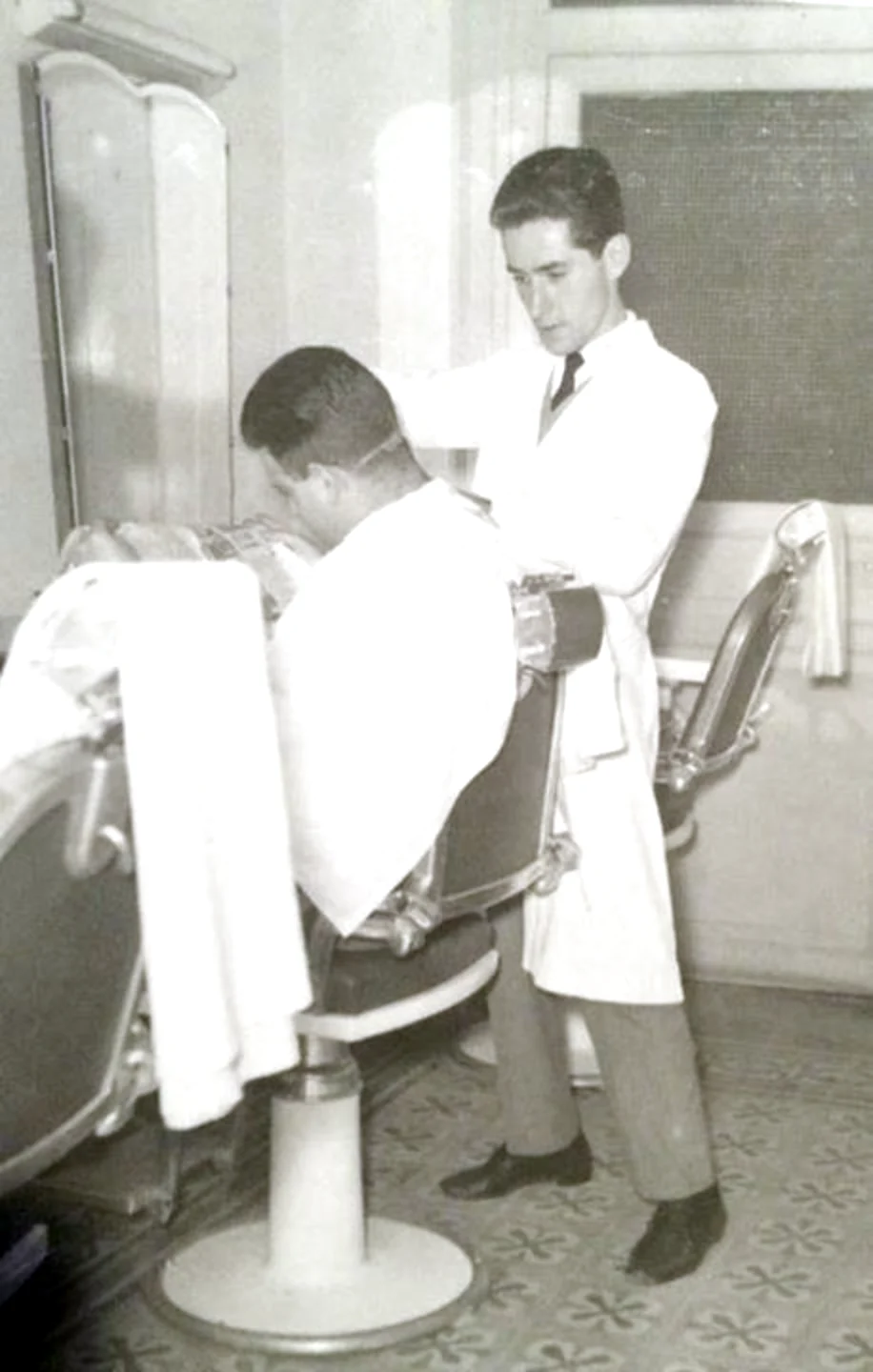 A young barber in a white coat and tie cutting a client's hair at Madison Men's Barbershop